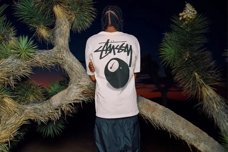 Mężczyzna w koszulce Stussy z grafiką kuli bilardowej 8 i logo Nike. Stussy to kultowa marka streetwearowa.