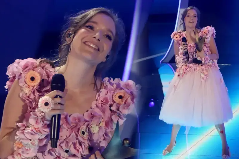 Zuzanna Irena Jurczak w kwiecistej sukni na scenie, trzyma mikrofon i uśmiecha się do publiczności.