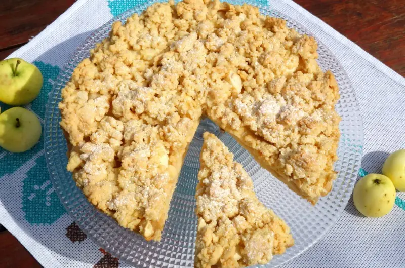 Szarlotka na kruchym cieście z kruszonką, obok jabłka, na talerzu.