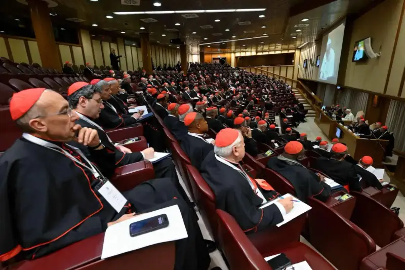 Wielu kardynałów w czerwonych biretach i czarnych szatach siedzi w amfiteatrze, słuchając przemówienia podczas ważnego konsystorza.