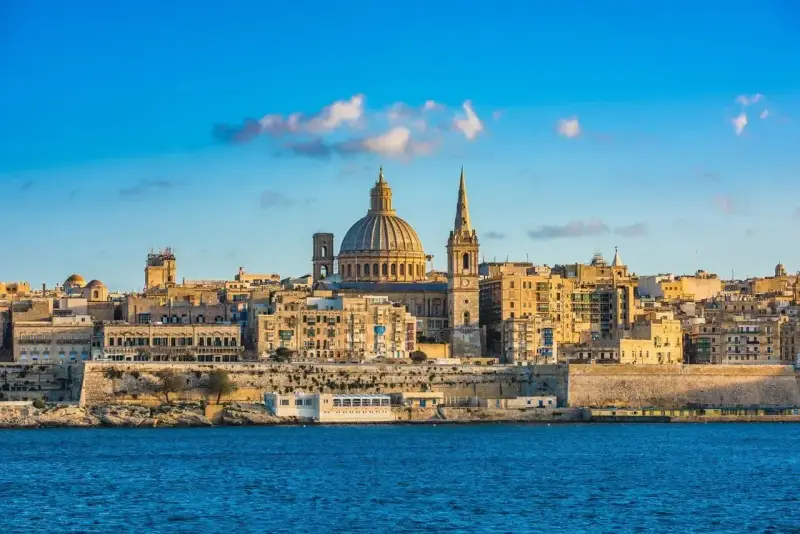 Malta, wyspa do kogo należy? Widok na Vallettę z kopułą i wieżą kościelną, otoczoną zabytkową architekturą, nad błękitnym morzem.