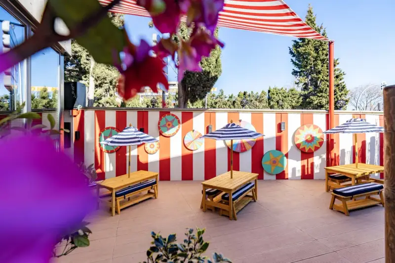 Terraza de restaurantes con zona infantil en Madrid, decorada con rayas rojas y blancas y coloridos adornos circulares. Mesas de madera con sombrillas a rayas invitan a disfrutar.
