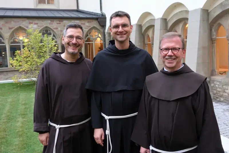 Drei Franziskaner Minoriten in braunen und schwarzen Kutten lächeln in die Kamera vor einer Klosterfassade.