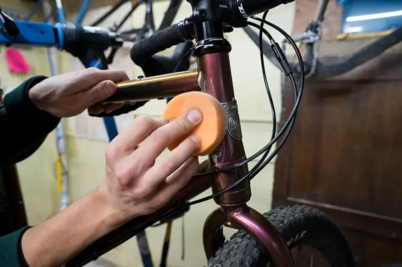 Hände polieren den Fahrradrahmen mit einem orangen Schwamm, um den Fahrradlack auszubessern.