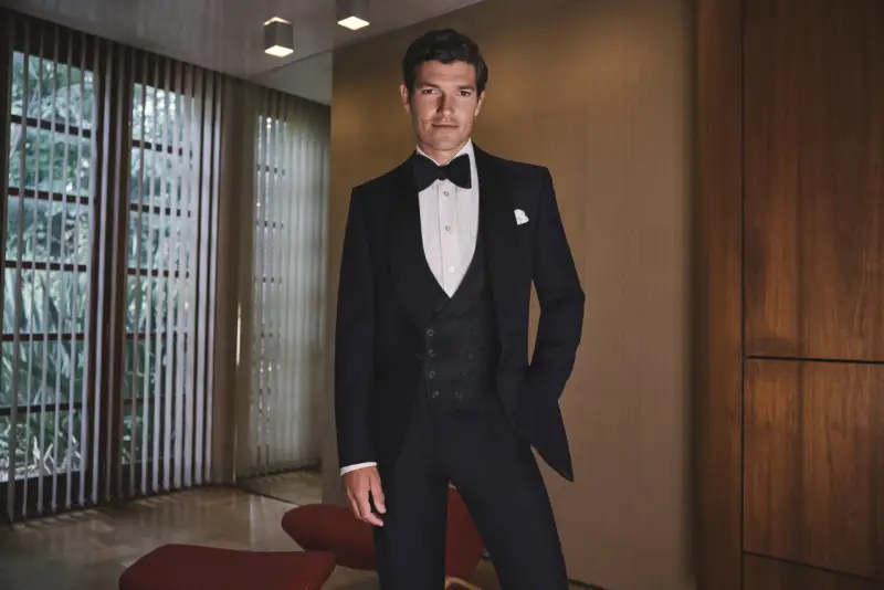 A man in a sharp black tie tuxedo, ready for a wedding. He stands confidently in a stylish room with large windows and wood paneling.