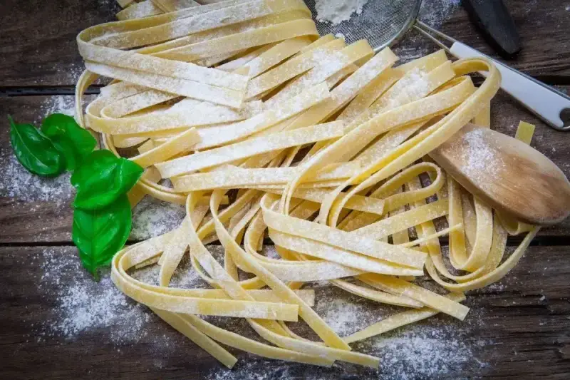 Świeży, domowy makaron bez glutenu, posypany mąką, z listkami bazylii i drewnianą łyżką.