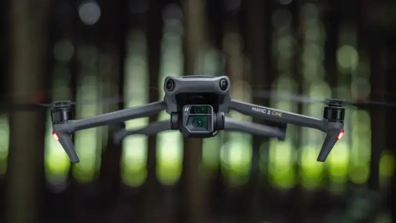 Szary dron Mavic 3 Cine zawieszony w powietrzu, z rozmytym zielonym lasem w tle.