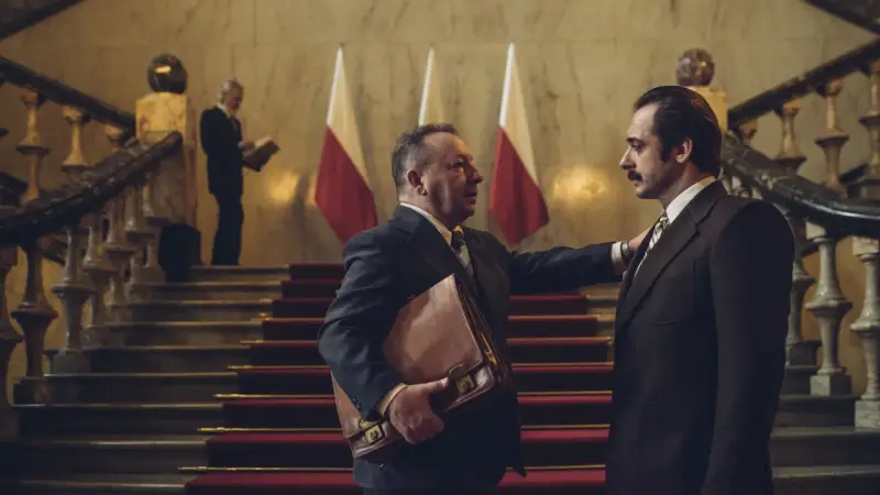 Hubert Niedziela, w garniturze, trzyma teczkę, rozmawiając z mężczyzną na schodach przed polskimi flagami.