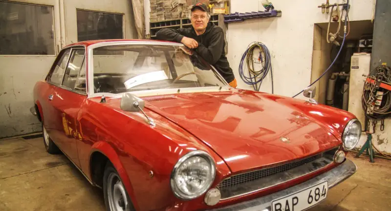 Man lutar sig mot en röd klassisk bil i garage. Lite rost syns på bilens skärmkanter.