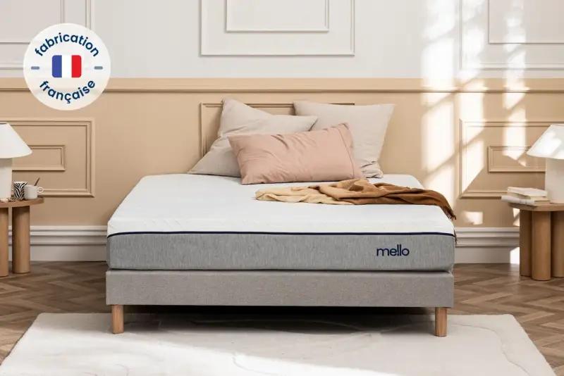 Matelas gris et blanc avec logo "mello", sur un sommier, avec des coussins et un plaid.