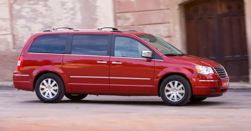 Czerwony Chrysler Grand Voyager w ruchu, z przyciemnianymi szybami i relingami dachowymi, przemierza ulicę obok kamiennej fasady.