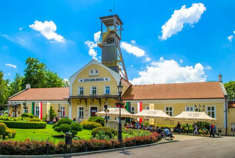 Wieliczka atrakcje: Szyb Daniłowicza, żółte budynki kopalni, zielony ogród i ludzie odpoczywający pod namiotami.