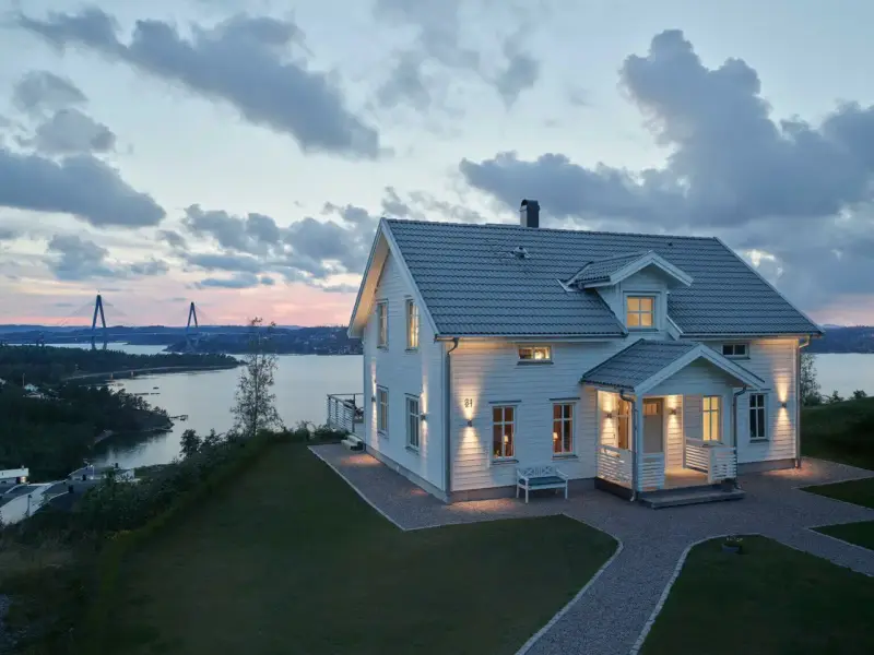 Vackert vitt hus med upplysta fönster och entré, perfekt hus entre inspiration. Vacker vy över vatten och bro i skymningen.