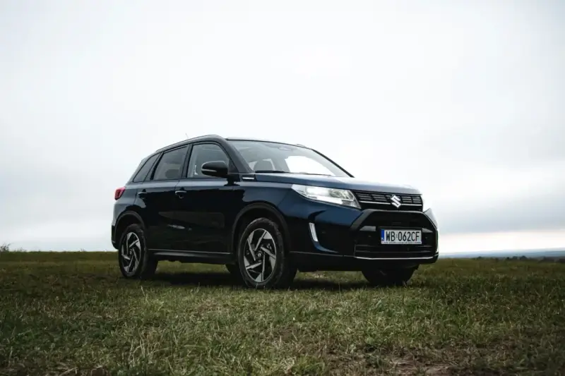 Ciemnoniebieski Suzuki Vitara Hybrid na tle zachmurzonego nieba, zaparkowany na trawiastym wzgórzu.