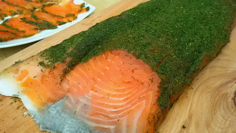 Un gran filete de salmón marinado casero cubierto de eneldo fresco. En el fondo, finas lonchas de salmón ya preparadas.