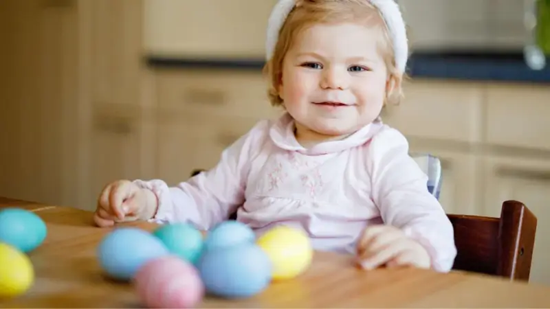 Kleines Mädchen mit Hasenohren und bunten Ostereiern. Sie sitzt am Tisch und schaut neugierig auf das **Baby Ei**.