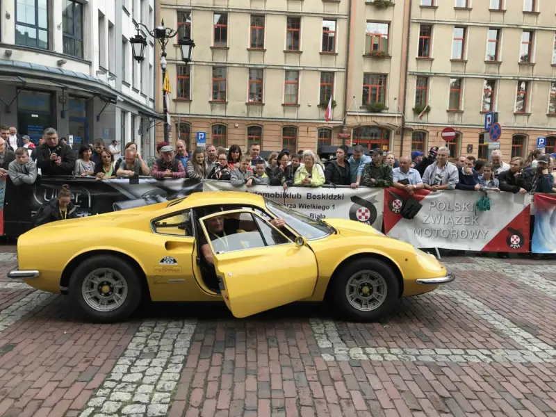 Żółte Ferrari Dino z otwartymi drzwiami, kierowca w środku, tłum widzów za barierką.