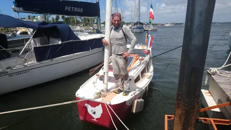 Yann Quenet, prêt pour l'aventure avec son baluchon, pose sur son voilier orné d'une poule.