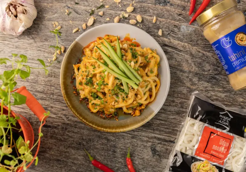 Makaron udon w sosie orzechowym, udekorowany ogórkiem i szczypiorkiem. Obok opakowanie makaronu udon mrożony czy suszony.