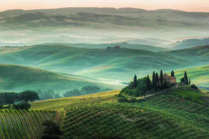 Un incantevole **punto panoramico Val d'Orcia** con dolci colline verdi, un casale in cima a una collina e filari di cipressi.