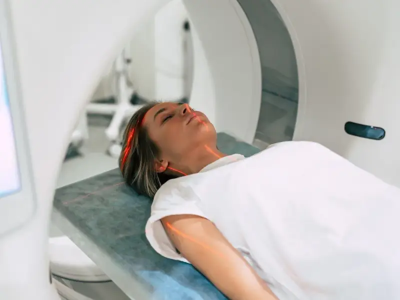 Kobieta leży na łóżku w aparacie do rezonansu magnetycznego. Czerwone linie lasera wskazują obszar badania. To właśnie mri co to za badanie?