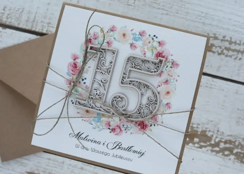 Elegancka kartka z napisem "15 rocznica ślubu Malwina i Bartłomiej" ozdobiona kwiatowym wianuszkiem i sznurkiem.
