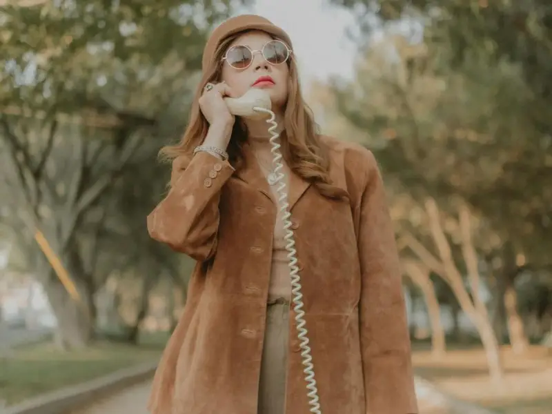 Kobieta w brązowej zamszowej kurtce damskiej rozmawia przez telefon w stylu retro.