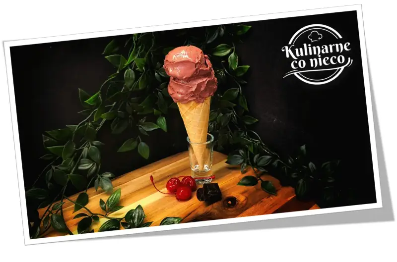 Dwa kule czekoladowych lodów białkowych w wafelku, ozdobione wiśniami i kawałkami gorzkiej czekolady.