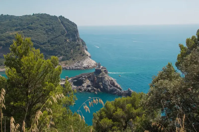 Vista panoramica del sentiero Riomaggiore Portovenere, con la chiesa di San Pietro su uno sperone roccioso e il mare azzurro.