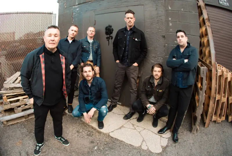 Les membres de Dropkick Murphys posent devant un mur sombre, certains accroupis, d'autres debout, prêts pour leur prochain concert.