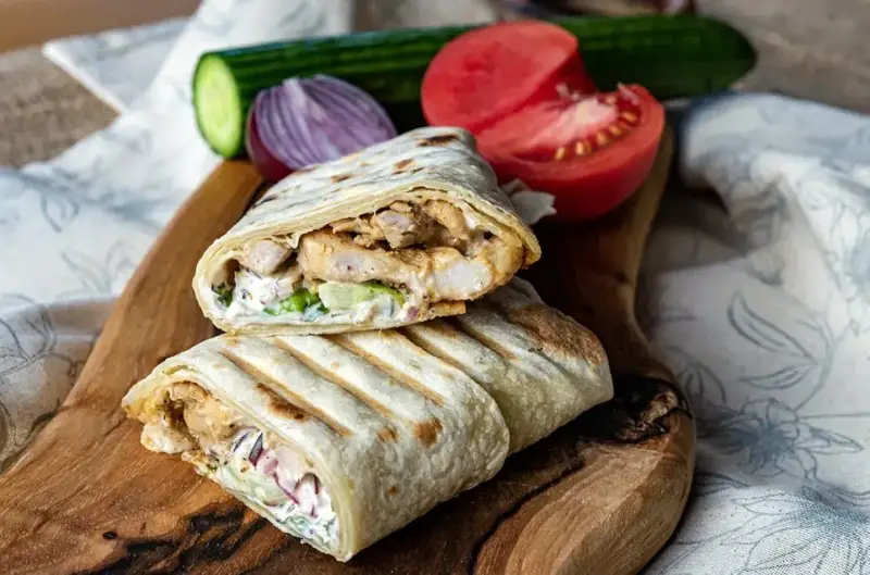 Pyszny, domowy kebab z kurczakiem, warzywami i sosem na desce z drewna. Obok pomidor, ogórek i cebula.
