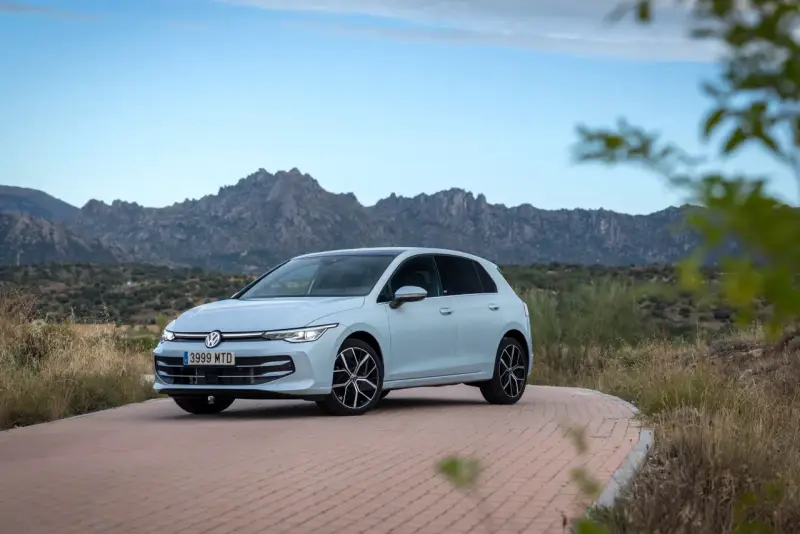 Un coche compacto Volkswagen Golf blanco circula por una carretera de adoquines con montañas rocosas al fondo.
