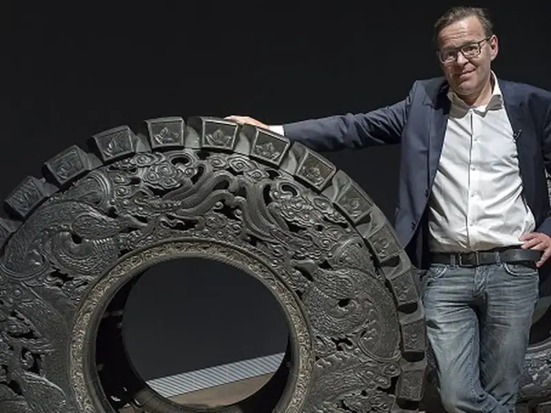 Wim Delvoye pose à côté d'une roue ornée de dragons, une œuvre d'art monumentale.
