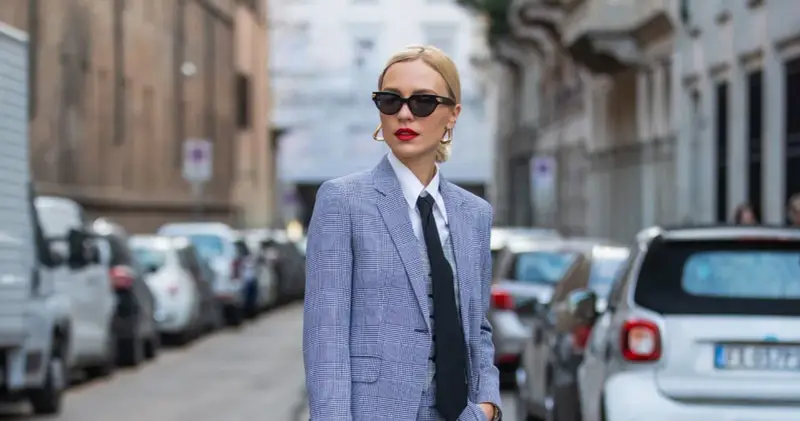Mujer con traje a cuadros, camisa blanca y corbata negra, luciendo un impecable tomboy style. Gafas de sol y labios rojos completan su look.