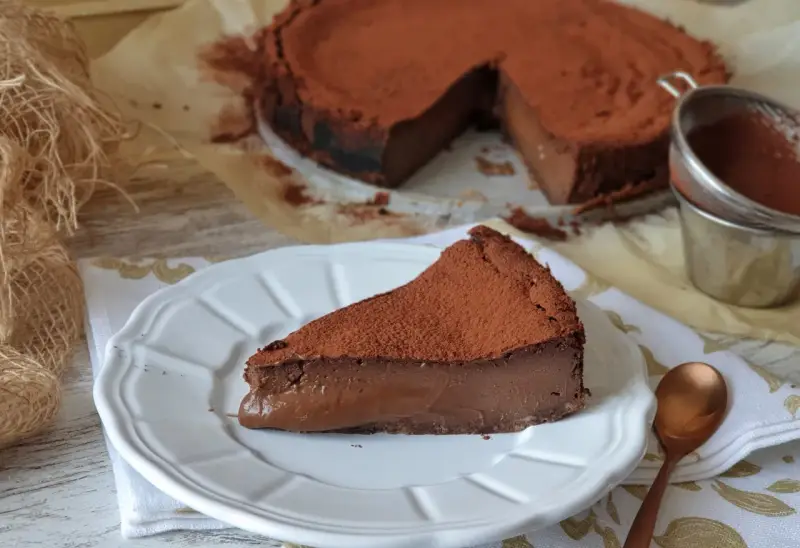 Una porción de tarta de chocolate, cubierta de cacao en polvo, reposa en un plato blanco. El resto de la tarta, también espolvoreada, se ve al fondo.