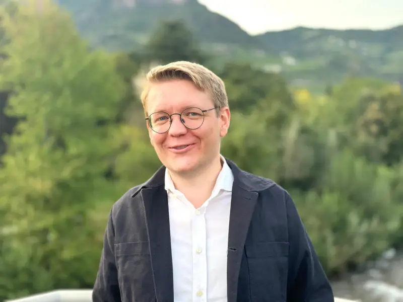 Ein Mann mit Brille lächelt in die Kamera. Er trägt ein weißes Hemd und eine dunkle Jacke. Im Hintergrund sind Bäume und Berge zu sehen. Er studiert Seelsorge.