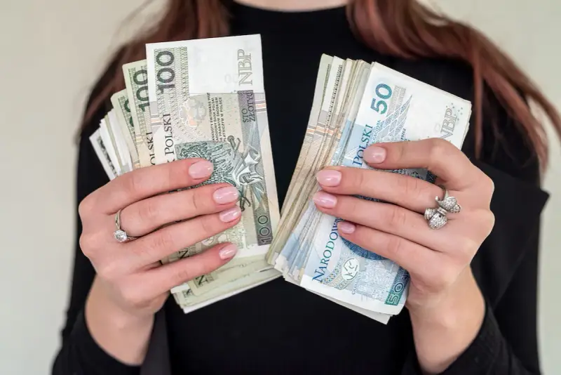 Dłonie z paznokciami w kolorze nude trzymają stosy banknotów 100 i 50 zł. To idealna jubileuszówka na prezent!