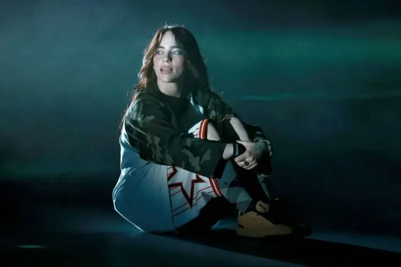 Billie Eilish w ciemnym pomieszczeniu, siedzi w pozie zamyślenia. Jej stylizacja nawiązuje do estetyki jej albumu.