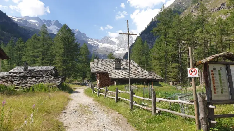 Sentiero di montagna con baite in pietra e tettoie, ideale per cogne escursioni facili. Montagne innevate sullo sfondo.