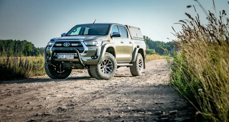 Złota toyota terenowa z wyciągarką i zabudową pędzi po polnej drodze, otoczona dziką roślinnością.