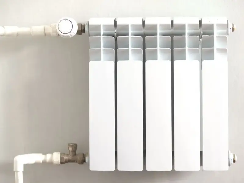 Biały grzejnik z termostatem, podłączony do instalacji rurowej. To efektywne ogrzewanie etażowe, które zapewnia komfort cieplny.