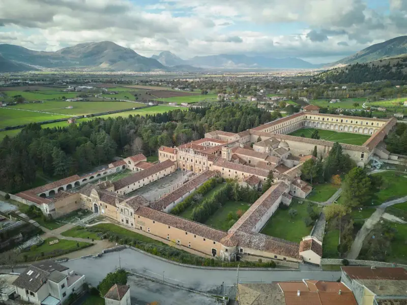 Vasta Certosa di Padula, un complesso monastico imponente con cortili verdi, circondato da alberi e montagne sullo sfondo.