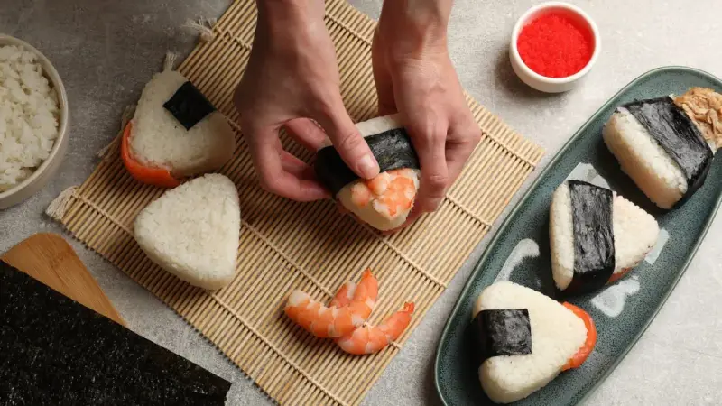 Ręce formują onigiri z krewetkami, tworząc pyszne japońskie jedzenie. Obok miska ryżu, nori i talerz gotowych przysmaków.