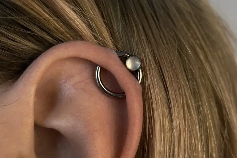 Oryginalny kolczyk orbital piercing z białym kamieniem na uchu, otoczony blond włosami.