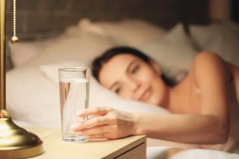 Une main attrape un verre d'eau posé à droite sur une table de chevet, une femme se réveille en arrière-plan.