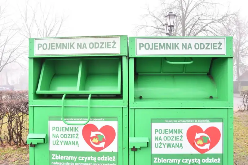 Dwa zielone kontenery na odzież używaną, z napisem "POJEMNIK NA ODZIEŻ UŻYWANĄ" i sercem z symbolami recyklingu.