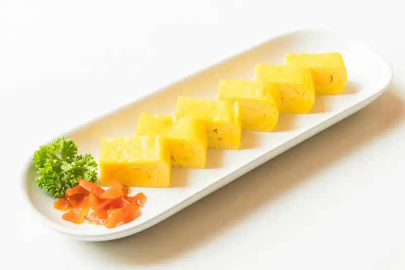 Sześć kawałków puszystego omletu tamago ułożonych na białej desce, z gałązką pietruszki i pomarańczowymi dodatkami.
