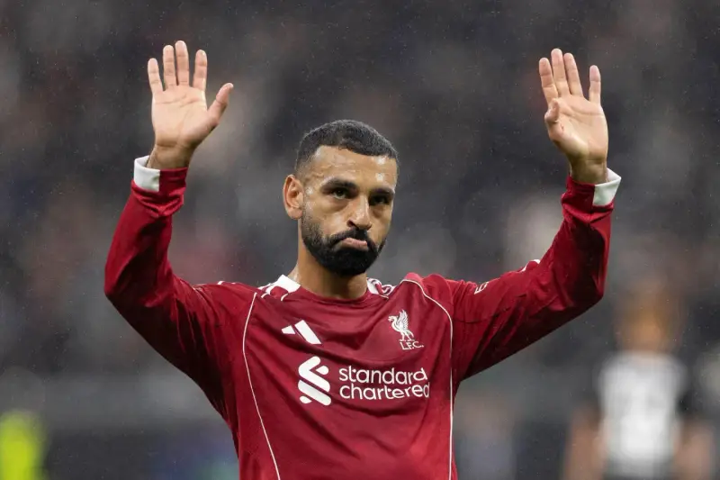 Mohamed Salah w czerwonej koszulce Liverpoolu, z uniesionymi dłońmi, pozdrawia kibiców. Widać logo klubu i sponsora.