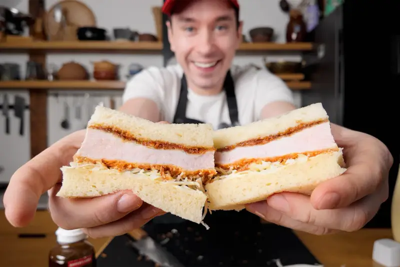 Uśmiechnięty mężczyzna prezentuje pyszne katsu sando z soczystym kotletem schabowym w panierce, delikatną kapustą i puszystym chlebem.