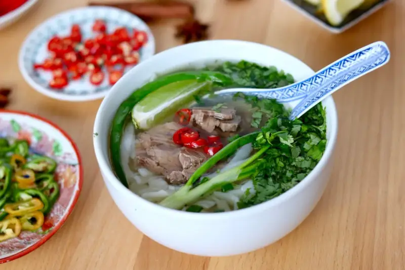 Aromatyczna zupa pho z wołowiną, makaronem ryżowym, świeżą kolendrą, chili i limonką. Obok talerzyki z dodatkami.